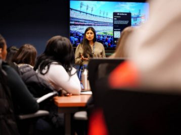 Detroit Tiger's employee giving a talk to students, with a picture of Comerica park on the TV on the wall behind her.